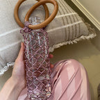 Pink woven water bottle carrier with wooden handles, held in hand beside striped pillow