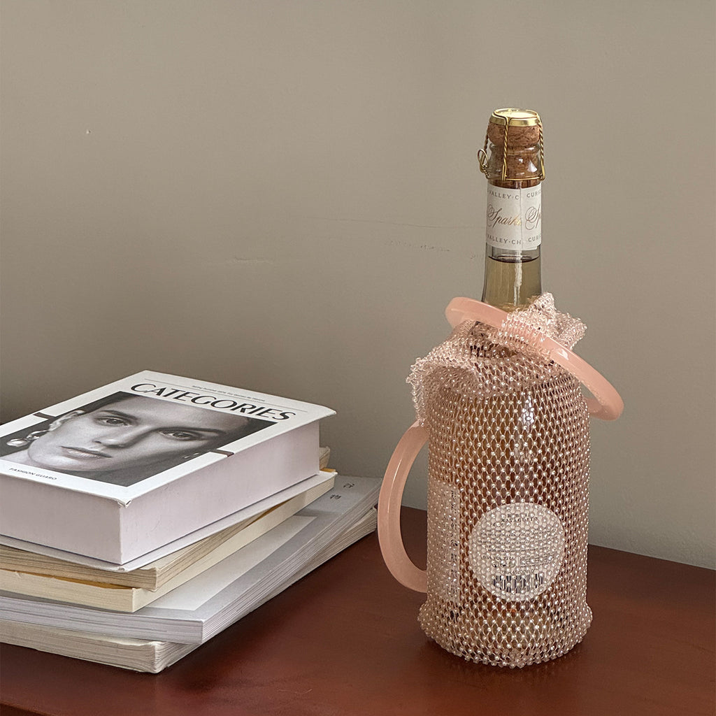 Pink rhinestone wine tote bag with handle, holding a sparkling wine bottle beside stacked books