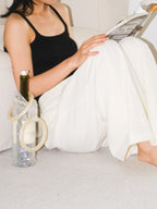 Sparkling rhinestone wine tote bag with cream handles, holding a bottle beside a woman reading on a couch.
