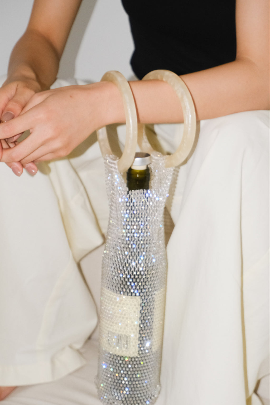 A close-up of a person wearing cream-colored wide-leg trousers and a black top, holding a wine bag covered in sparkling silver rhinestones. The bag features two thick, cream-colored circular handles and a bottle cap closure at the neck.