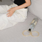 A woman wearing a black camisole and white pants sits on a light gray carpet next to a crystal wine bag bottle filled with glitter and two beige rings.