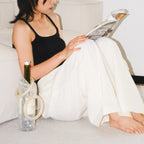 A woman sits barefoot on a light carpet in a minimalist room, wearing a black tank top and white wide-leg pants while reading a magazine. A clear textured bottle with a gold cap and a crystal wine bag rest beside her.